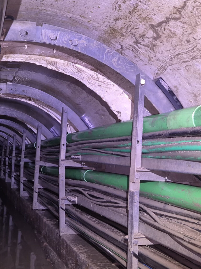 Underground tunnel with bundled green and gray pipes along the curved concrete ceiling and metal supports; a wet floor.
